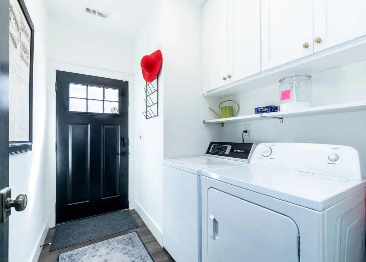 A practical nook featuring a white washer and dryer, black front door, and a shelf with household essentials. The space is functional yet inviting, suggesting convenience for guests, especially families or longer-stay groups.