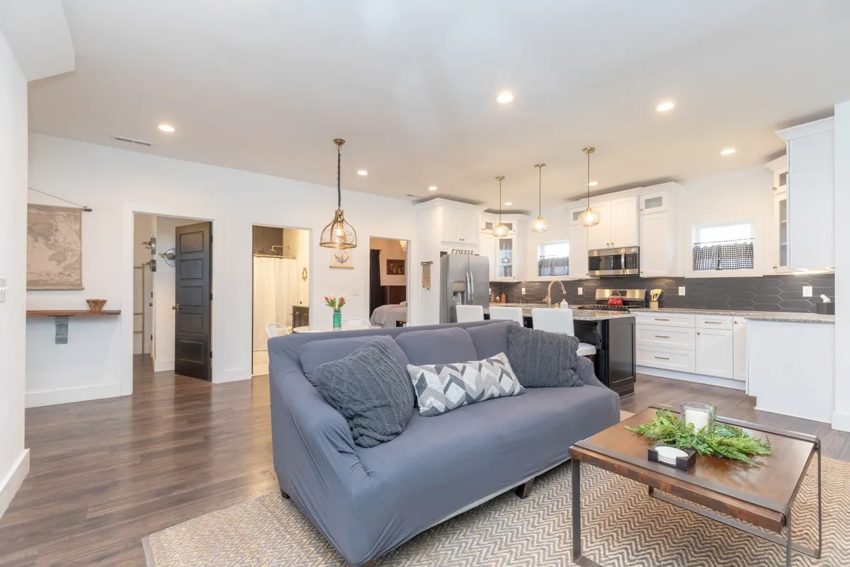 This image features an open-concept living area that seamlessly blends a comfortable sofa, a spacious kitchen island, and a dining area illuminated by pendant lights. The layout suggests ample space for socializing, dining, and group gatherings, with a warm, inviting atmosphere ideal for entertaining friends or family.