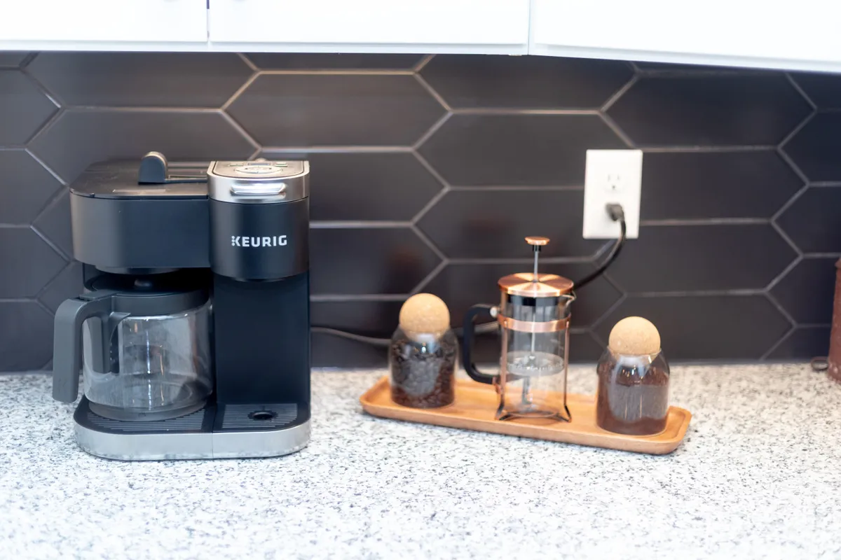 A cozy kitchen coffee station with a Keurig, French press, glass jars, and a granite countertop, inviting guests to make their perfect cup of coffee and chat.