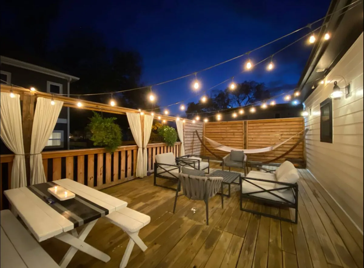 The image showcases the exterior of the Nashville vacation home at dusk, with warm string lights casting a cozy glow over a spacious patio. There's a hammock inviting guests to relax, an outdoor dining table set for candlelit meals, and plenty of seating for socializing or simply enjoying the starry Tennessee sky.