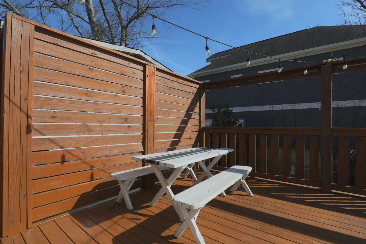 The backyard deck features a wooden patio with a white picnic table under string lights, suggesting space for outdoor dining, play, and family gatherings in a relaxed, festive atmosphere. The string lights and picnic table hint at fun al fresco meals and celebrations.