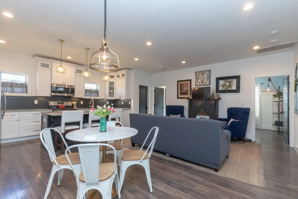 This scene focuses on a dining table adorned with flowers, flanked by blue armchairs, with the kitchen island and a fireplace in the background. The arrangement encourages socializing, shared meals, and relaxation in a stylish, inviting setting. The view includes both the dining and living spaces, reinforcing connectivity.