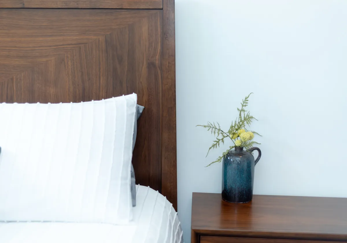 A tranquil bedroom with a wooden bed frame, crisp white bedding, and a decorative vase with flowers on the nightstand. Soft daylight illuminates the peaceful setting, making it inviting for rest or quiet moments.